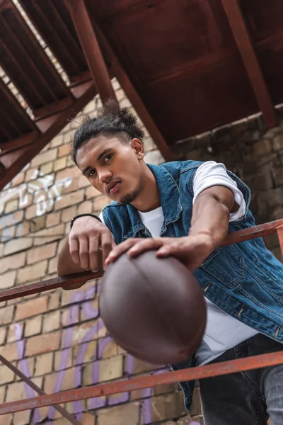 Vista de ángulo bajo del hombre de raza mixta mostrando pelota de rugby en la calle urbana - foto de stock