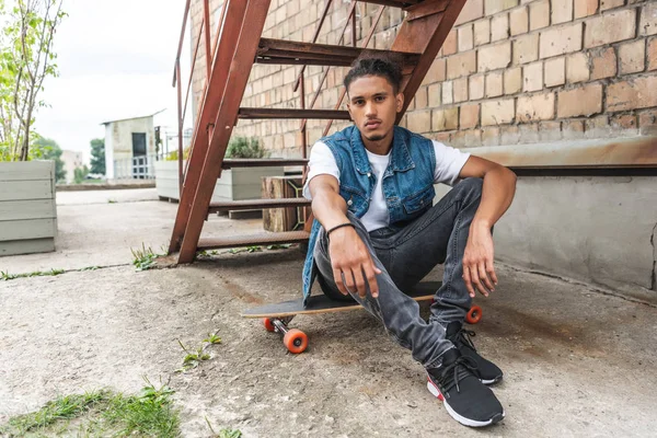 Junger stylischer Mischlingsrennfahrer sitzt auf Skateboard und blickt in die Kamera auf der Stadtstraße — Stockfoto