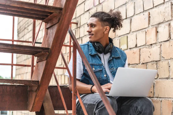 Concentré mixte homme freelance race dans les écouteurs travaillant avec ordinateur portable et regardant loin de la rue — Photo de stock