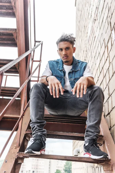 Low angle view of serious stylish mixed race man sitting on stairs at urban street — Stock Photo