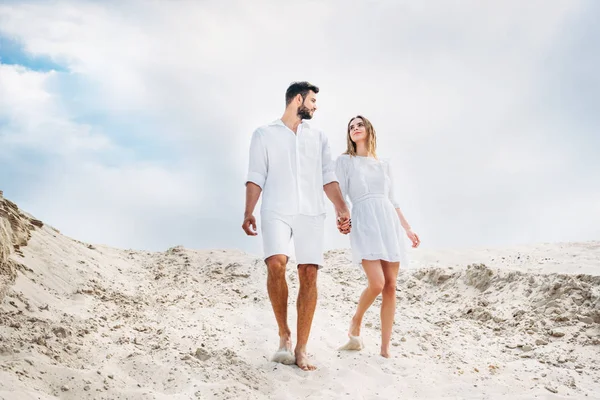Beautiful young couple in white clothes walking by sand with bare feet — Stock Photo