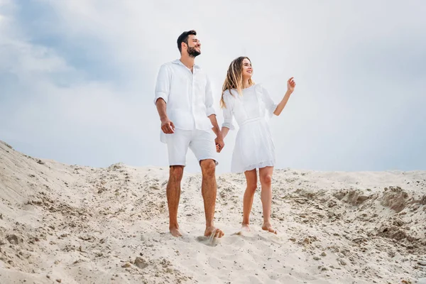 Happy young couple in white clothes walking by sand with bare feet — Stock Photo