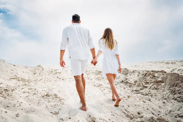 Vista posteriore della giovane coppia in abiti bianchi che cammina dalla sabbia con i piedi nudi — Foto stock
