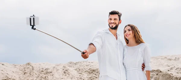 Smiling young couple taking selfie with monopod and smartphone in desert — Stock Photo