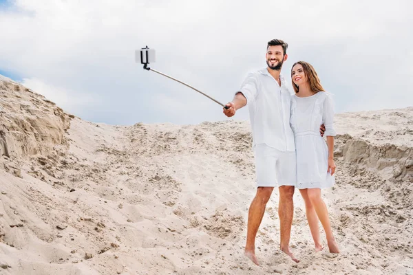 Beautiful young couple in white clothes taking selfie with monopod and smartphone in desert — Stock Photo