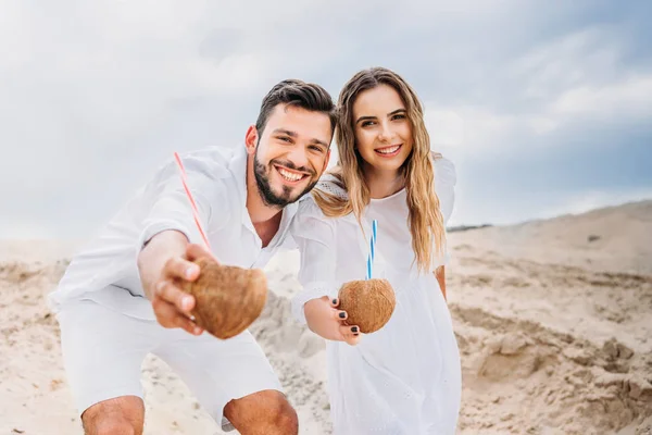 Счастливая молодая пара в белом с кокосовыми коктейлями, смотрящая в камеру — стоковое фото