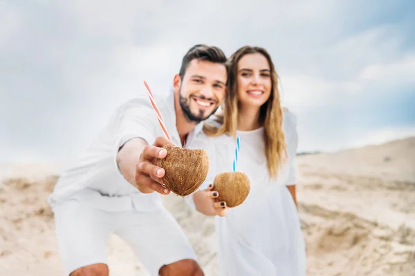 Sonriente joven pareja en blanco con cocteles de coco mirando a la cámara - foto de stock