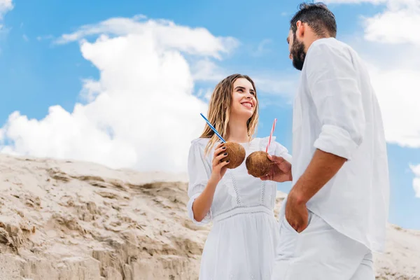 Vista inferior de feliz pareja joven en blanco con cócteles de coco en frente del cielo azul — Stock Photo