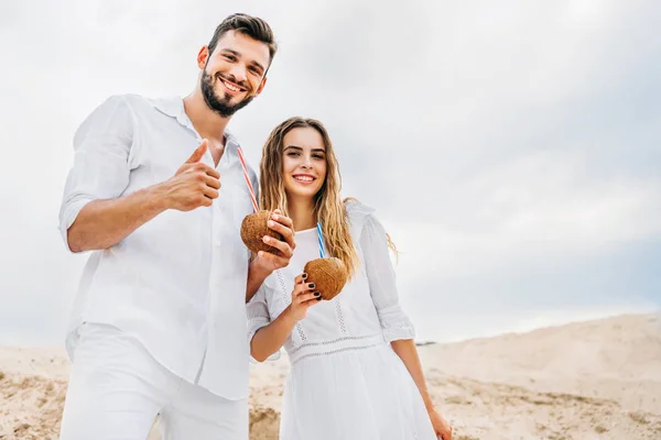 Happy young couple in white with coconut cocktails looking at camera — Stock Photo