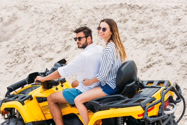 Beautiful young couple in sunglasses riding all-terrain vehicle in desert — Stock Photo
