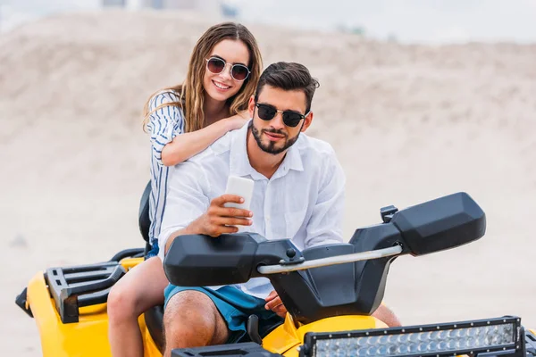 Happy young couple taking selfie while sitting on ATV in desert — Stock Photo