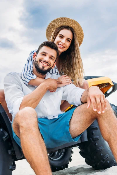 Blick von unten auf ein glückliches junges Paar, das auf einem ATV vor bewölktem Himmel sitzt — Stockfoto