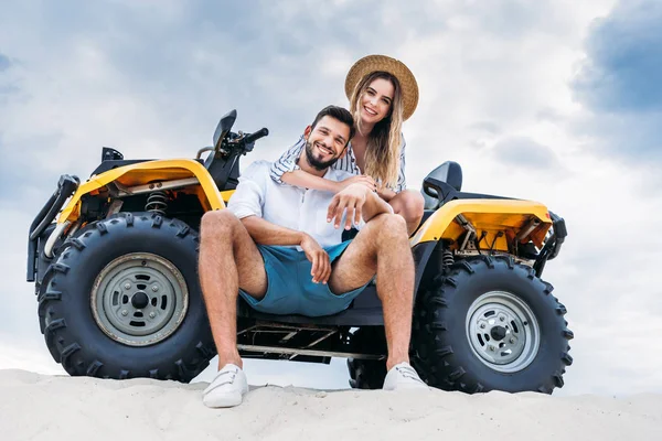 Bottom view of happy young couple sitting on ATV on sandy dune in front of cloudy sky — Stock Photo