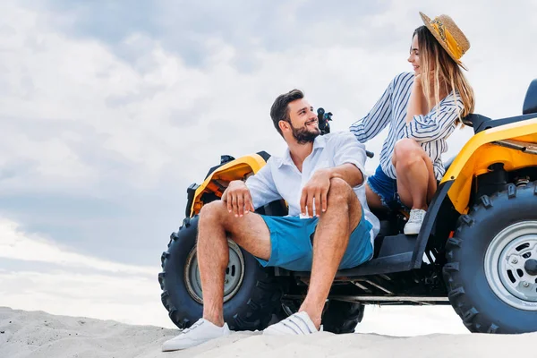 Feliz jovem casal sentado no ATV na duna arenosa na frente do céu nublado — Fotografia de Stock