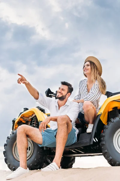 Schönes junges Paar sitzt auf ATV auf Sanddüne und zeigt irgendwo hin — Stockfoto