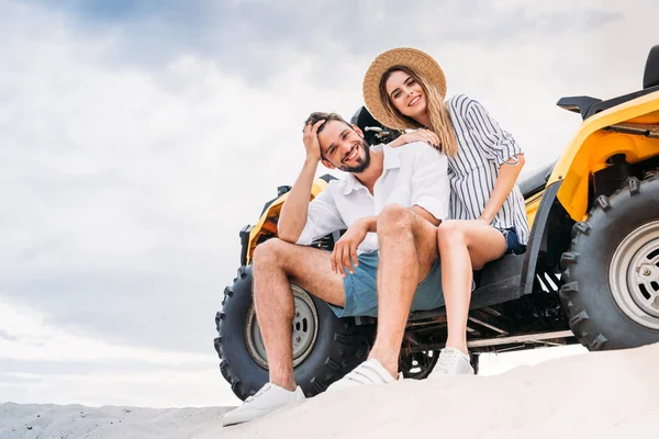 Hermosa joven pareja sentado en ATV en duna de arena y mirando a la cámara - foto de stock