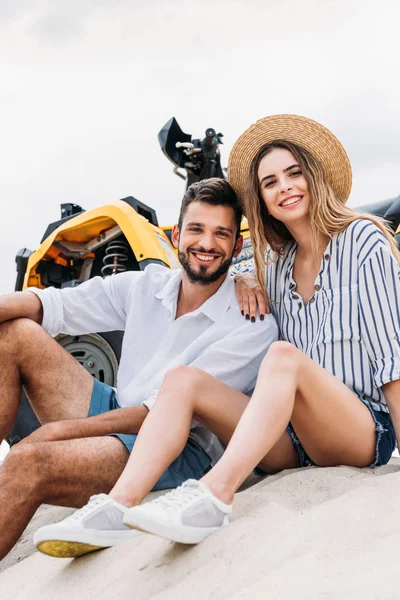 Feliz jovem casal sentado perto de ATV na duna arenosa e olhando para a câmera — Fotografia de Stock