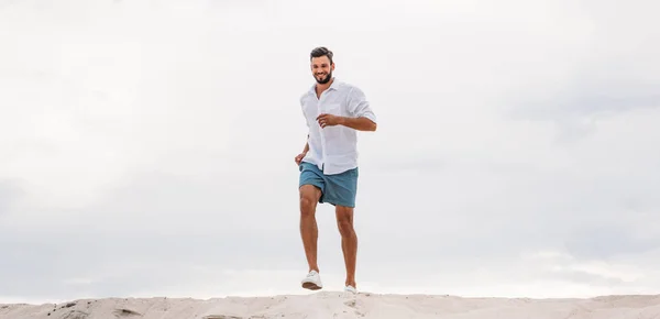 Beau jeune homme courant sur une dune de sable devant un ciel nuageux — Photo de stock