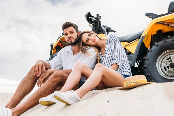Vista inferior de la hermosa pareja joven sentada cerca de ATV en duna de arena y mirando a la cámara - foto de stock