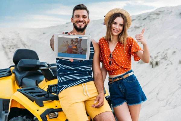 Feliz jovem casal com ATV mostrando tablet digital com site couchsurfing na tela e polegar no deserto — Fotografia de Stock