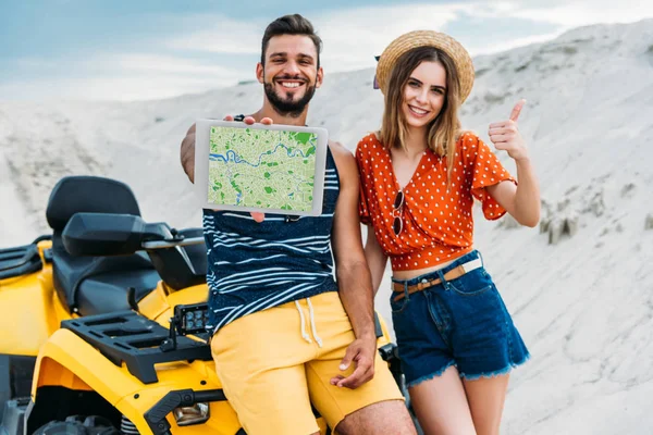 Schönes junges Paar mit ATV zeigt digitales Tablet mit Navigations-Website auf dem Bildschirm und Daumen hoch in der Wüste — Stockfoto