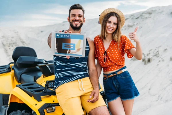 Beautiful young couple with atv showing digital tablet with fly ticket booking website on screen and thumb up at desert — Stock Photo