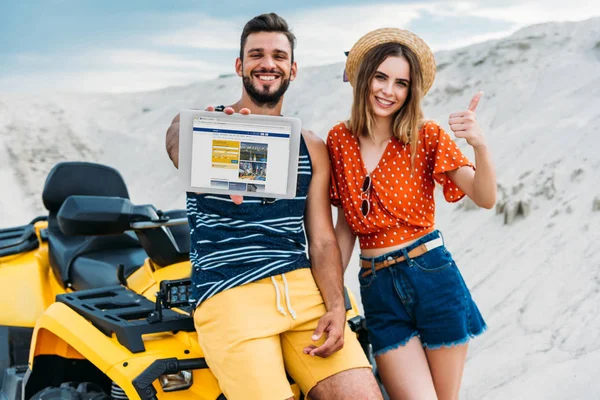 Belo jovem casal com ATV mostrando tablet digital com site de reserva na tela e polegar no deserto — Fotografia de Stock