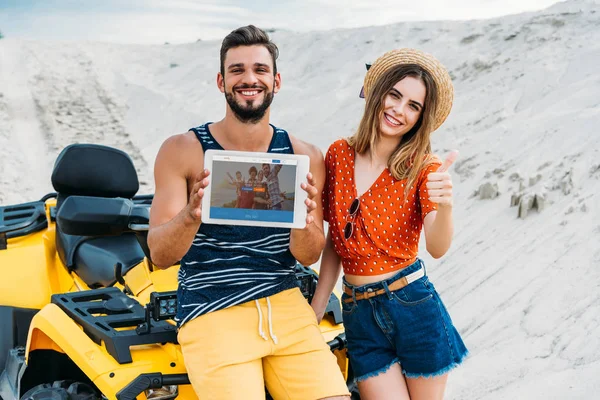 Schönes junges Paar mit ATV zeigt digitales Tablet mit Couchsurfing-Website auf dem Bildschirm und Daumen hoch in der Wüste — Stockfoto