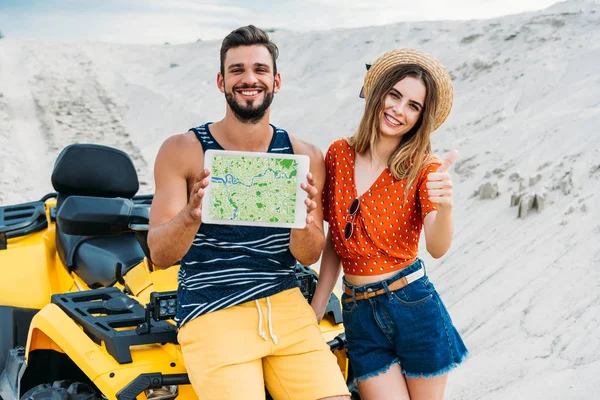 Beau jeune couple avec VTT montrant tablette numérique avec carte à l'écran et pouce vers le haut au désert — Photo de stock