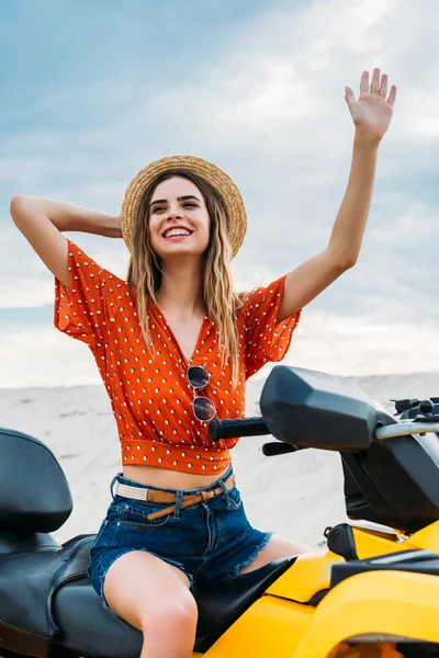 Smiling young woman sitting on all-terrain vehicle in desert waving hand — Stock Photo