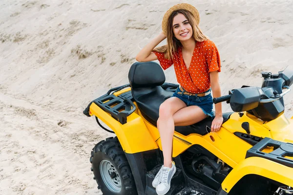 Vue grand angle de jeune femme heureuse assise sur un véhicule tout-terrain sur du sable — Photo de stock