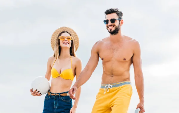 Hermosa pareja joven en ropa de playa con pelota de voleibol en frente del cielo nublado - foto de stock