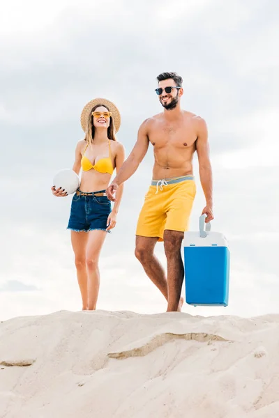 Beau jeune couple en vêtements de plage avec ballon de volley-ball et réfrigérateur portable marchant sur le sable — Photo de stock