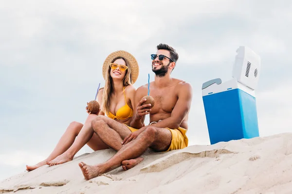 Jovem casal sorridente com coquetéis de coco relaxando na areia na frente do céu nublado — Fotografia de Stock