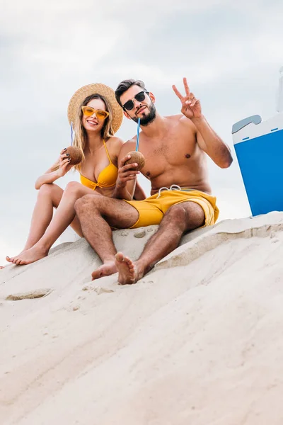 Vista inferior de pareja joven con cocteles de coco sentados en la arena frente al cielo nublado y mostrando gesto de paz — Stock Photo