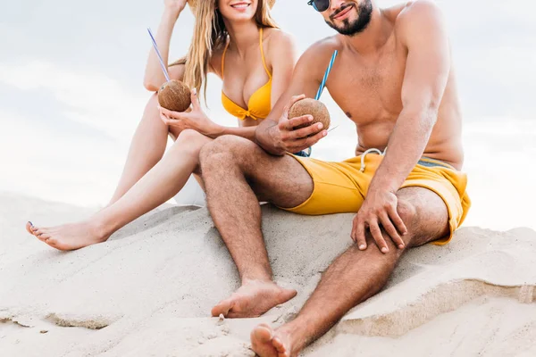 Tiro recortado de pareja joven con cócteles de coco sentado en la arena frente al cielo nublado - foto de stock
