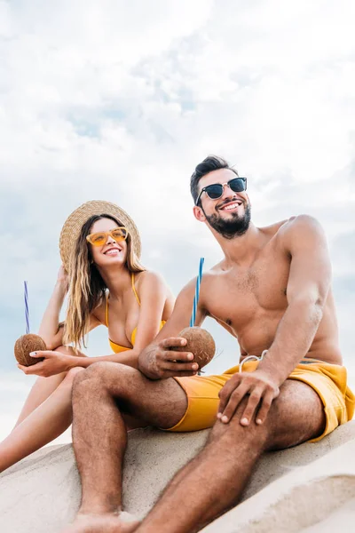 Bottom view of young couple with coconut cocktails sitting on sand in front of cloudy sky — Stock Photo