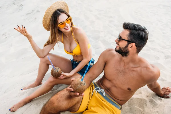 Vista de ángulo alto de la pareja joven con cócteles de coco sentado en la arena - foto de stock