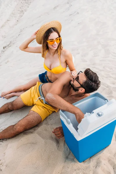 Vue grand angle du jeune couple avec réfrigérateur portable assis sur le sable — Photo de stock