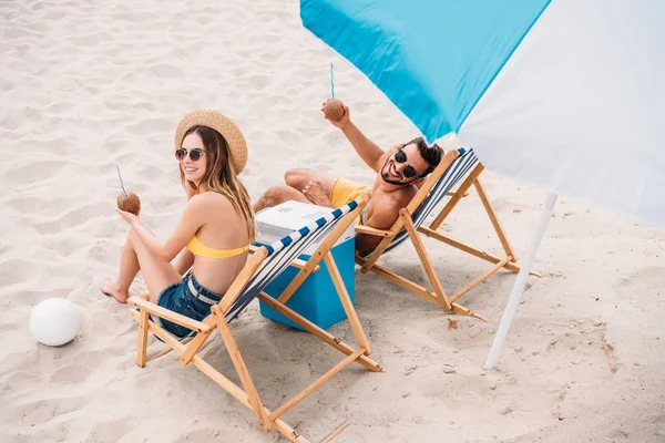 Vue grand angle du jeune couple heureux avec des cocktails de noix de coco relaxant dans des chaises longues sur la plage de sable — Photo de stock