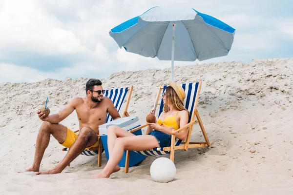 Belo jovem casal com coquetéis de coco relaxante em espreguiçadeiras na praia de areia — Fotografia de Stock