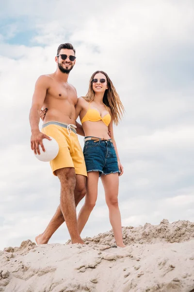 Belo jovem casal com bola de vôlei andando por duna arenosa na frente do céu nublado — Fotografia de Stock