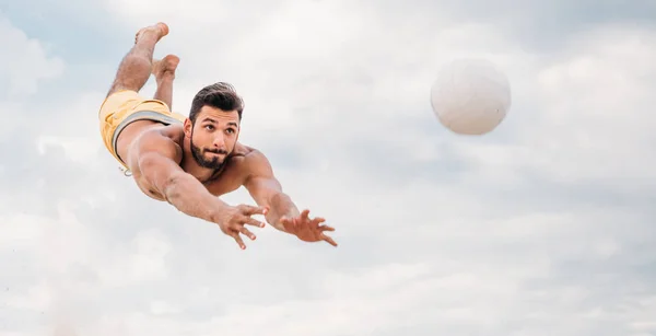 Jovem bonito pulando para bola enquanto joga vôlei de praia na frente do céu nublado — Fotografia de Stock
