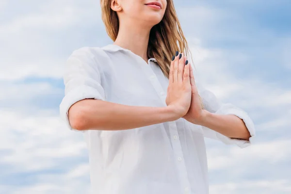 Обрезанный снимок молодой женщины медитирующей руками, делающей мудру намасте перед облачным небом — стоковое фото