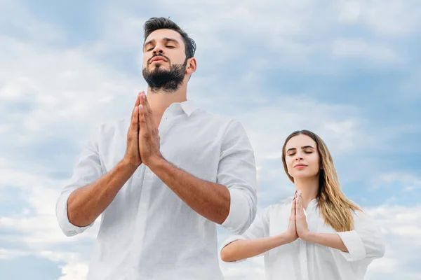 Blick von unten auf ein junges Paar, das meditiert und namaste mudra vor bewölktem Himmel macht — Stockfoto