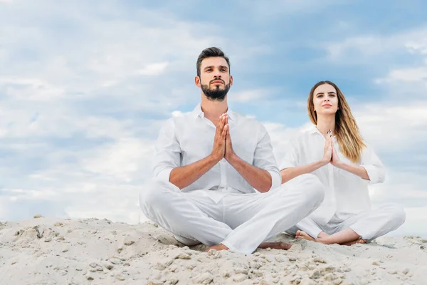 Hermosa pareja joven practicando yoga mientras se sienta en una duna de arena en pose de loto (padmasana ) - foto de stock