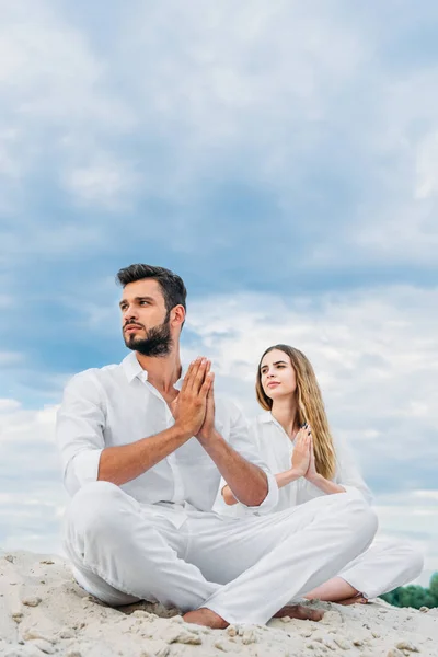 Beautiful young couple practicing yoga while sitting on sandy dune in lotus pose (padmasana) and looking away — Stock Photo