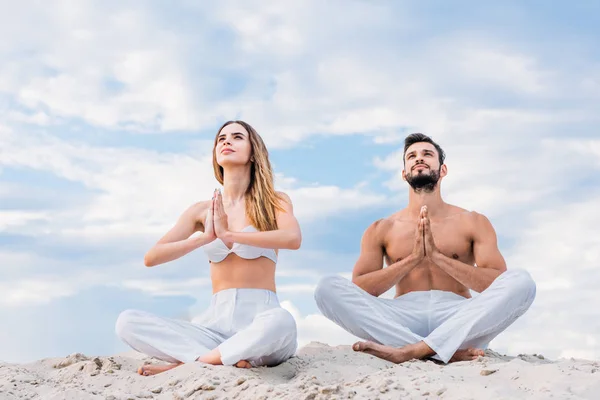 Fit young couple meditating while sitting on sandy dune in lotus pose — Stock Photo