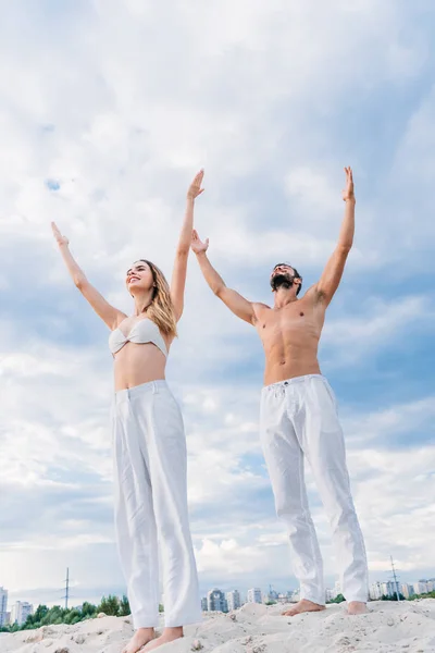 Vista inferior de la pareja joven en forma con los brazos levantados meditando bajo el cielo nublado - foto de stock