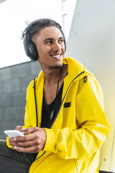 Feliz hombre de raza mixta en auriculares escuchando música con teléfono inteligente y mirando hacia la calle de la ciudad - foto de stock